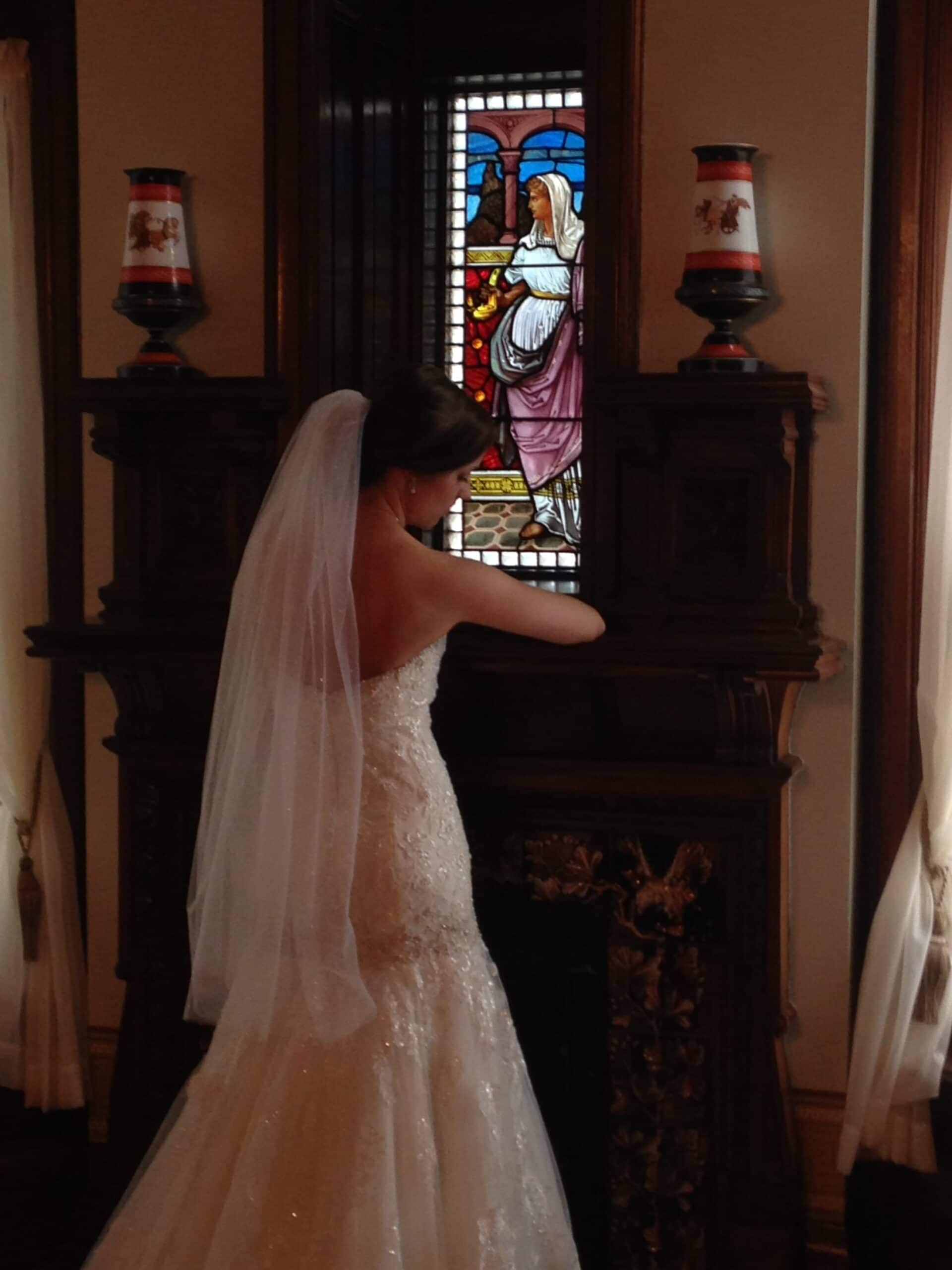 A bride in a white gown stands near a stained glass window, adorned with decorative lamps.