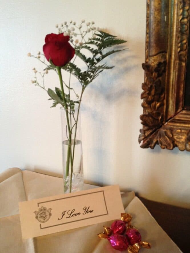 A single red rose in a glass vase alongside a "I Love You" card and chocolates on a table.