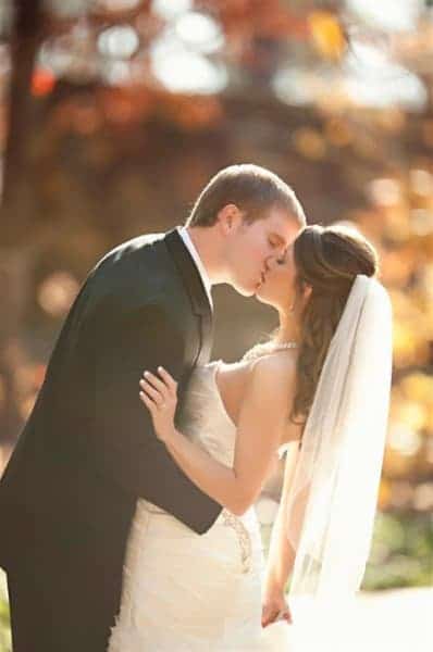 A couple in formal wedding attire shares a kiss outdoors, surrounded by soft, golden light.