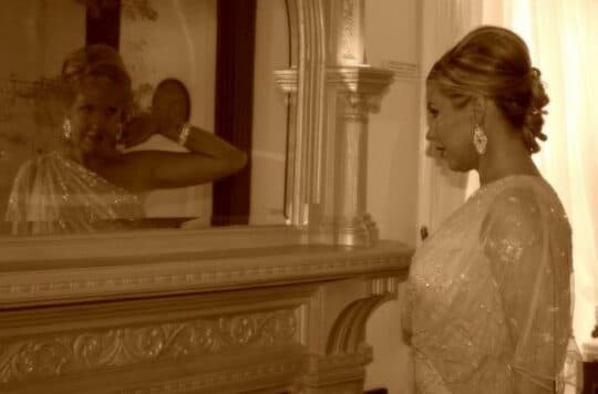 A woman in an elegant outfit gazes at her reflection in a mirror.