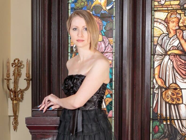 A woman in a black strapless dress poses near colorful stained glass windows.