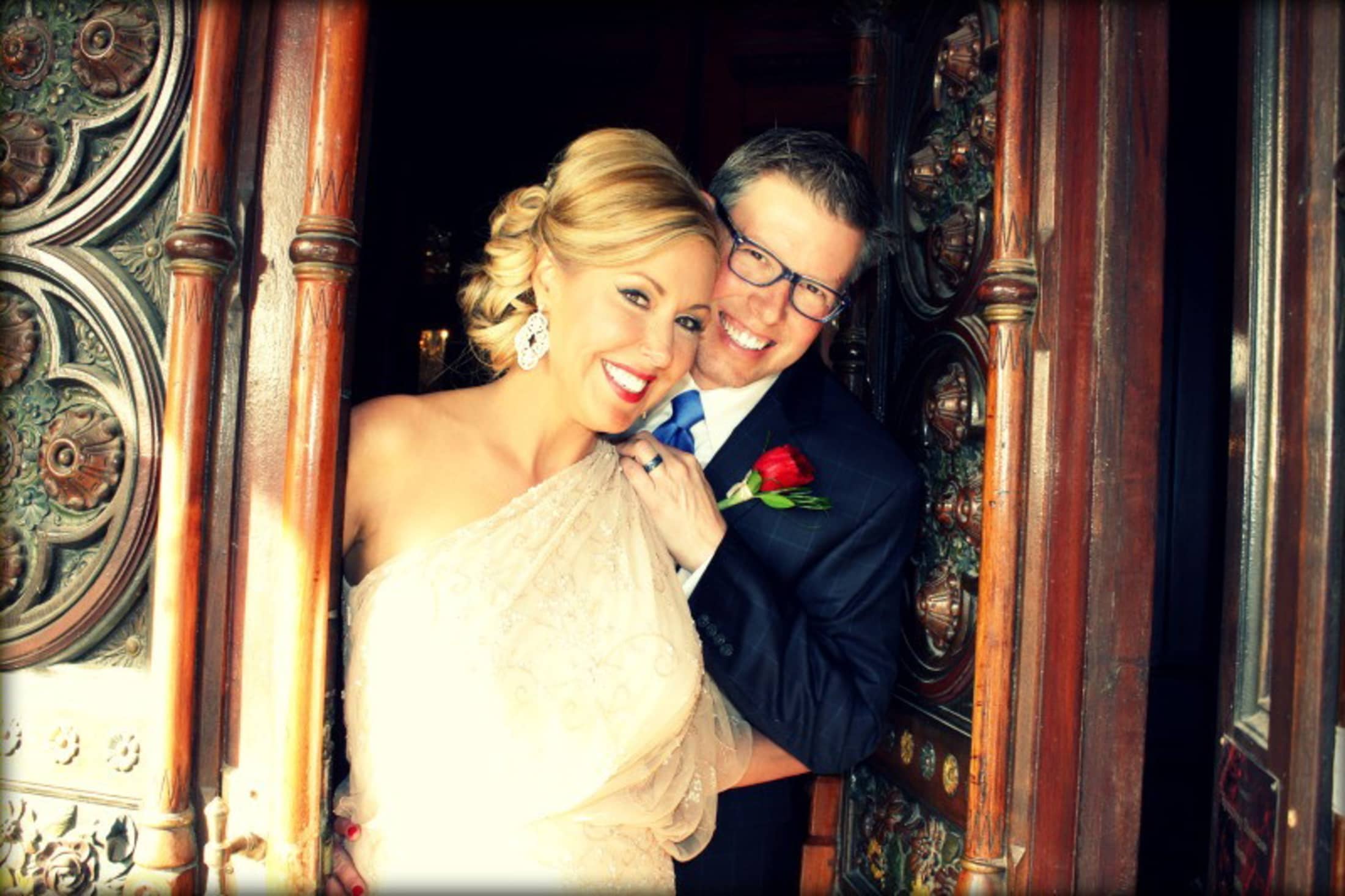 A smiling couple poses together at an ornate door, with the woman in a strapless dress and the man in a suit holding a rose.