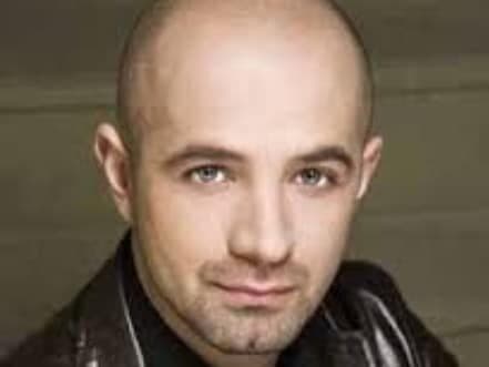 A close-up portrait of a man with a shaved head and a slight smile, wearing a dark leather jacket.