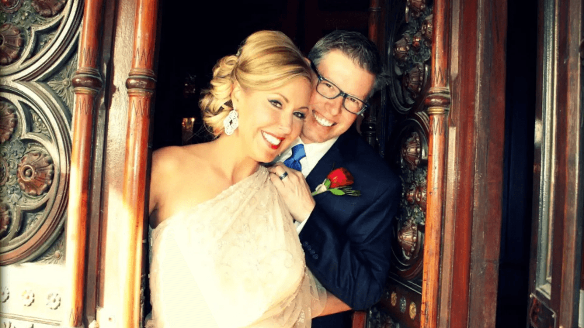 A smiling couple stands together at an ornate door, wearing formal attire, with the woman in a light dress and the man in a suit.