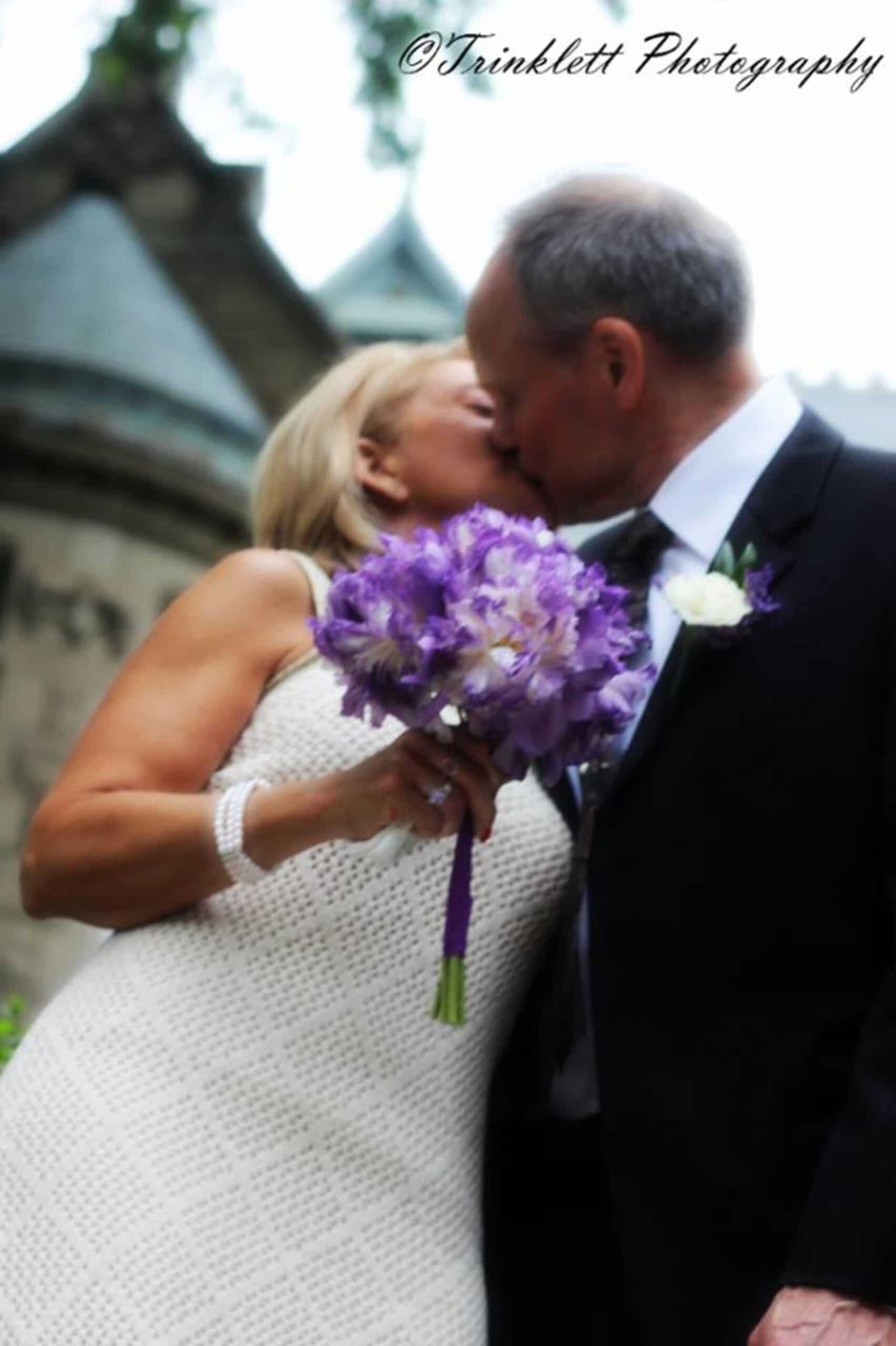A couple sharing a kiss while holding a bouquet of purple flowers.
