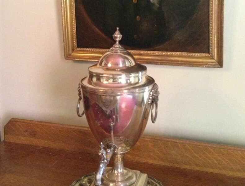 A silver urn sits on a wooden surface beneath an ornate portrait.