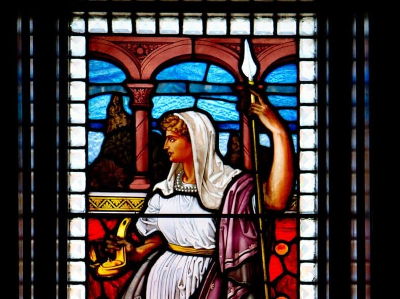 Stained glass window depicting a woman holding a spear and a small object, wearing a white dress and a veil.