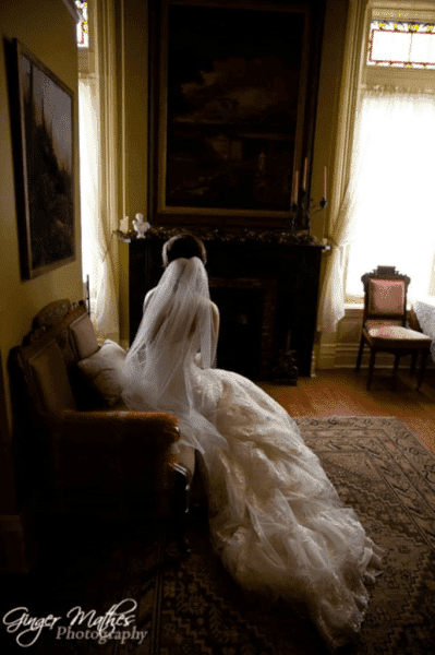 A bride in a flowing wedding gown sits thoughtfully on a vintage sofa in a warmly lit room.