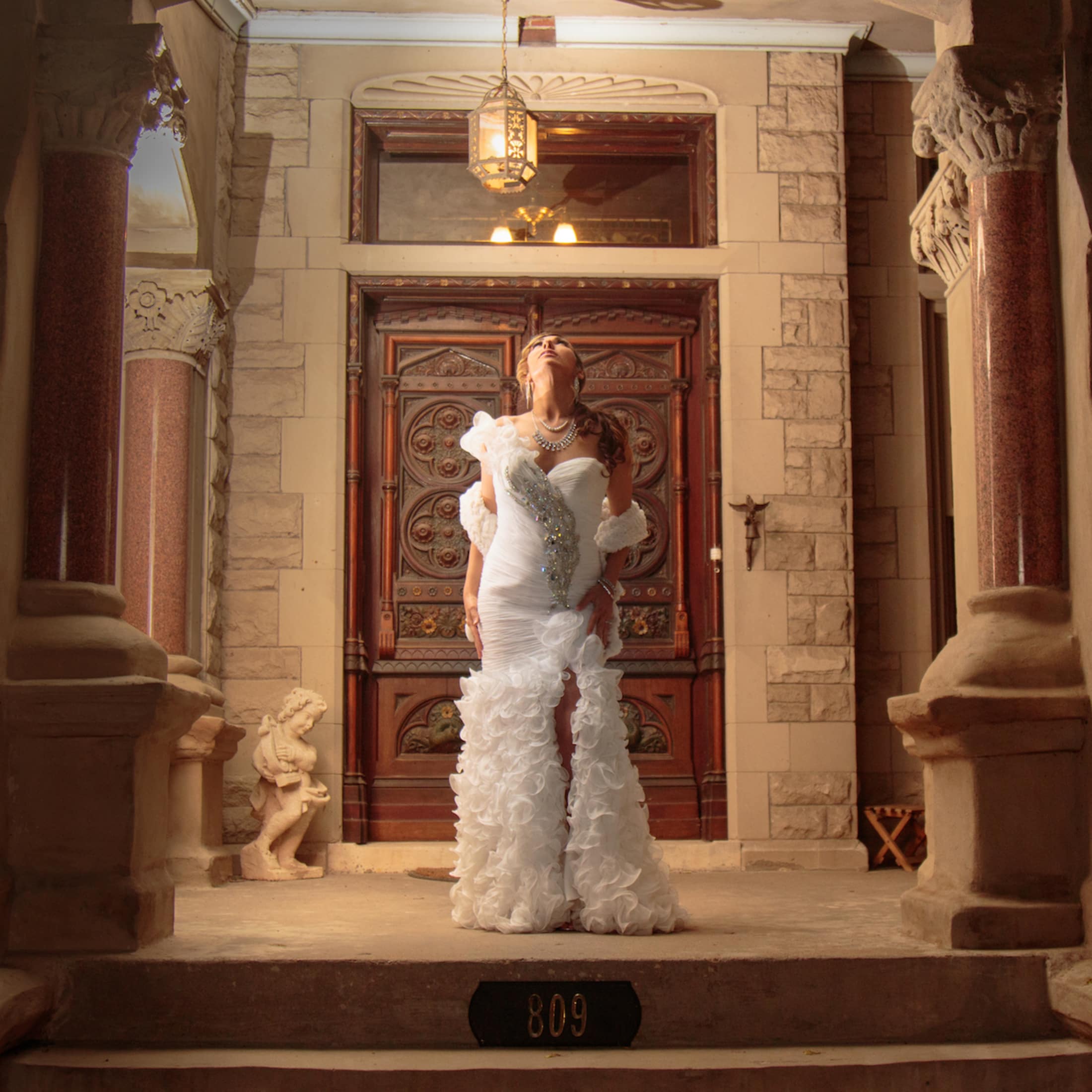 A woman in an ornate white dress stands confidently at the entrance of a vintage home.