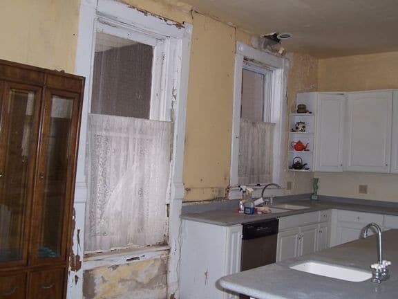 Kitchen with peeling paint, two windows with lace curtains, and white cabinets.