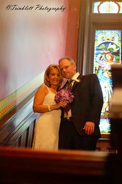 A couple poses together on a staircase, with a bouquet of flowers and colorful stained glass in the background.