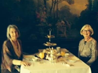 Two women are seated at a table set for tea in a warmly-lit room with a large painting in the background.