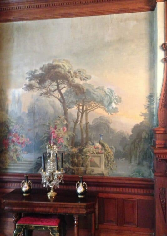 A vintage mural featuring trees and flowers adorns a wooden wall near an ornate table and decorative vases.
