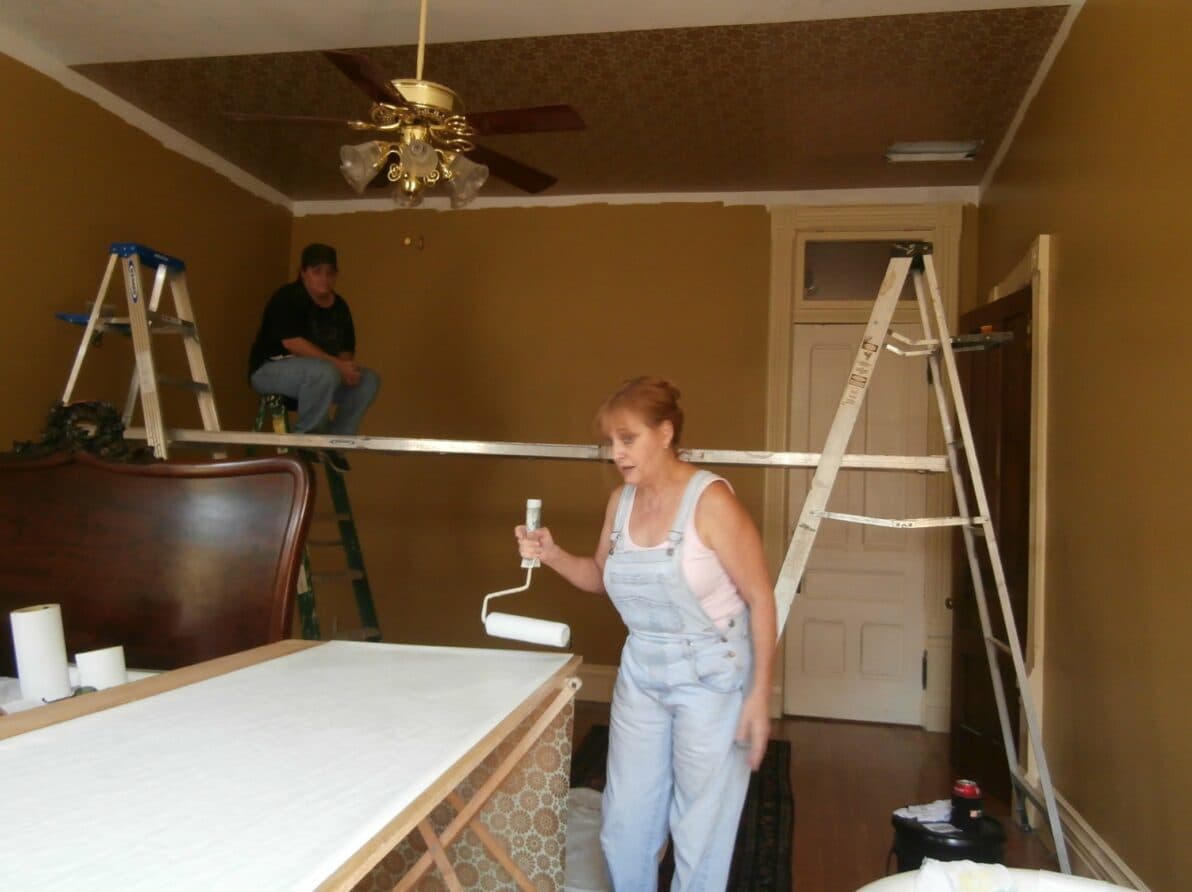 A woman paints the ceiling while a man sits on a ladder in a room undergoing renovation.
