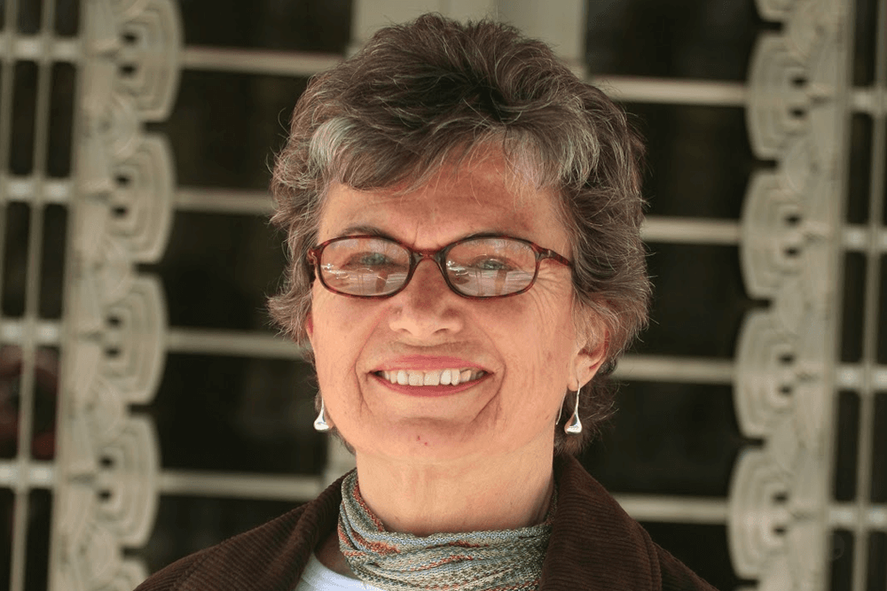 An older woman with short gray hair and glasses smiles in front of a decorative metal screen.